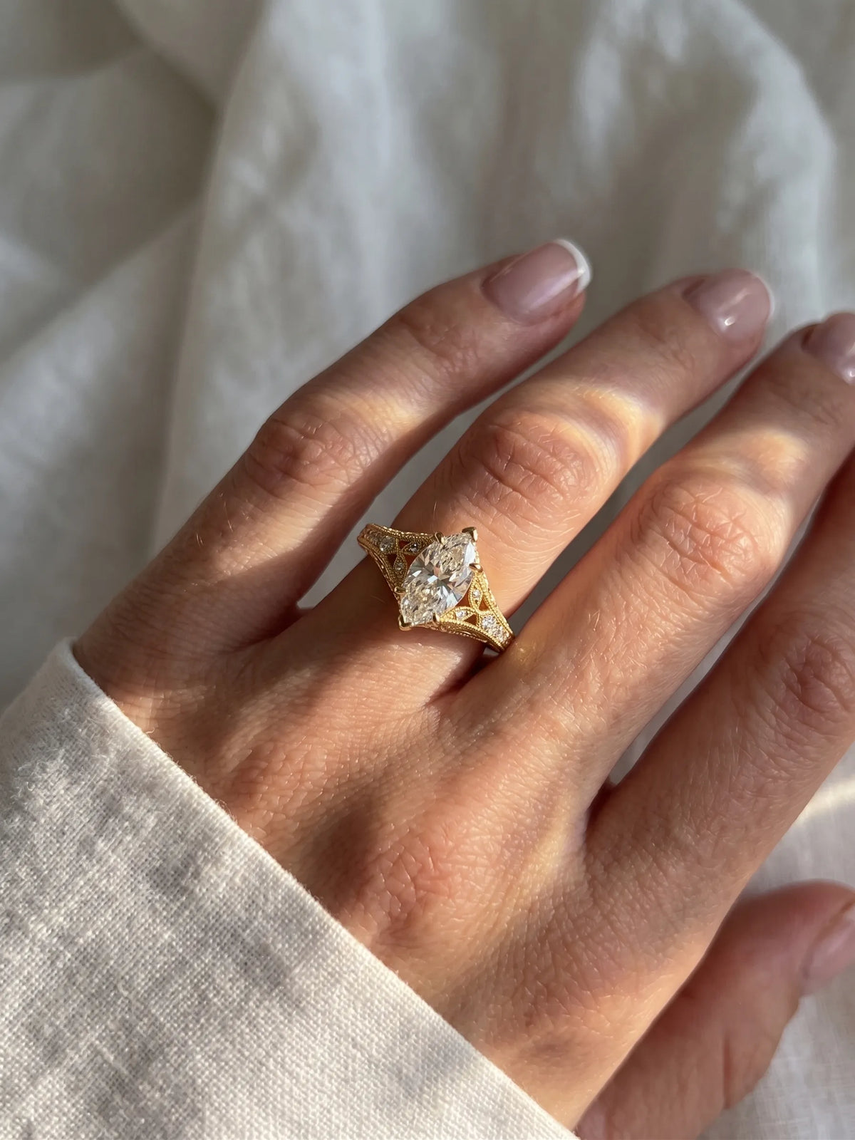 A gold ring with a marquise-cut clear gemstone and small side stones, worn on a hand resting against a soft white fabric background.