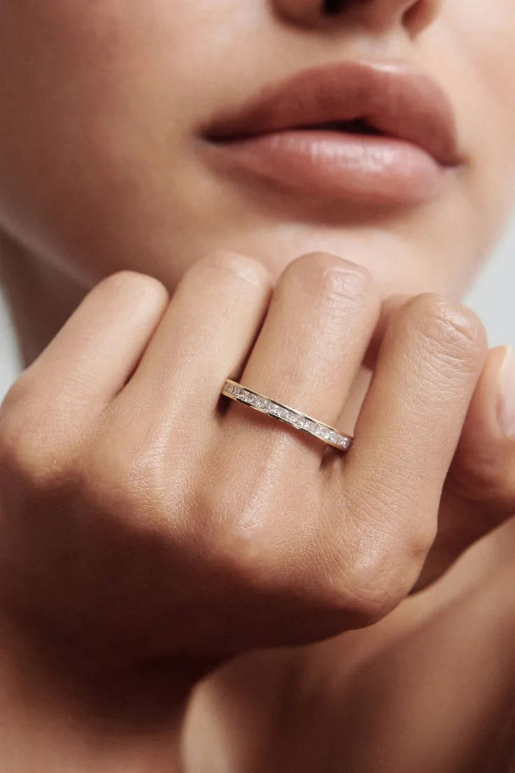 A close-up of a model's hand wearing a delicate silver band ring with a row of small clear stones, set against a soft-focus background of the model's face.