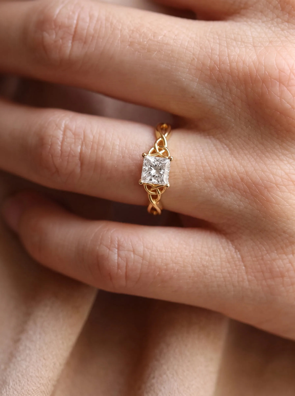 A gold ring with a princess-cut clear gemstone worn on a finger, shown in a close-up lifestyle shot against skin tones.