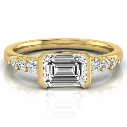 Emerald cut lab grown diamond engagement ring featuring a horizontal east-west bezel-set center diamond with graduated round side diamonds in a yellow gold tone.