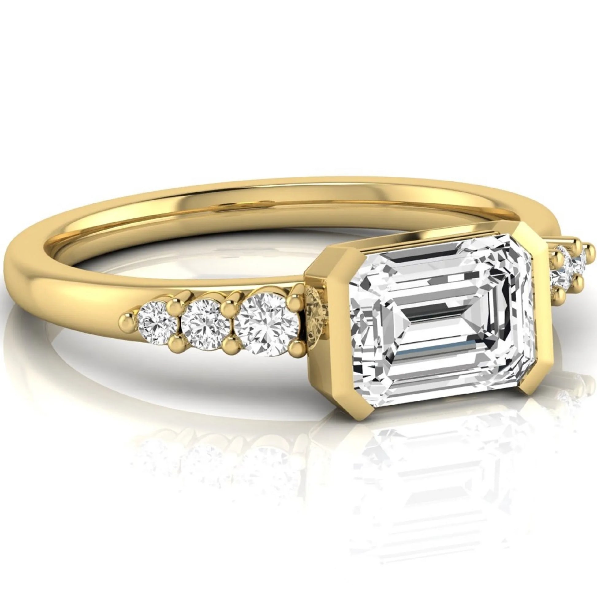 Emerald cut lab grown diamond engagement ring featuring a horizontal east-west bezel-set center diamond with graduated round side diamonds in a yellow gold tone.