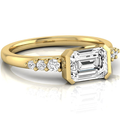 Emerald cut lab grown diamond engagement ring featuring a horizontal east-west bezel-set center diamond with graduated round side diamonds in a yellow gold tone.